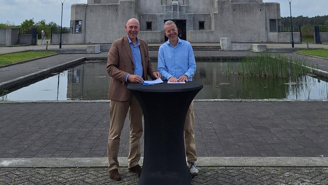 Twee bestuurders staan lachend achter een statafel bij Radio Kootwijk, na het tekenen van de intentieverklaring.